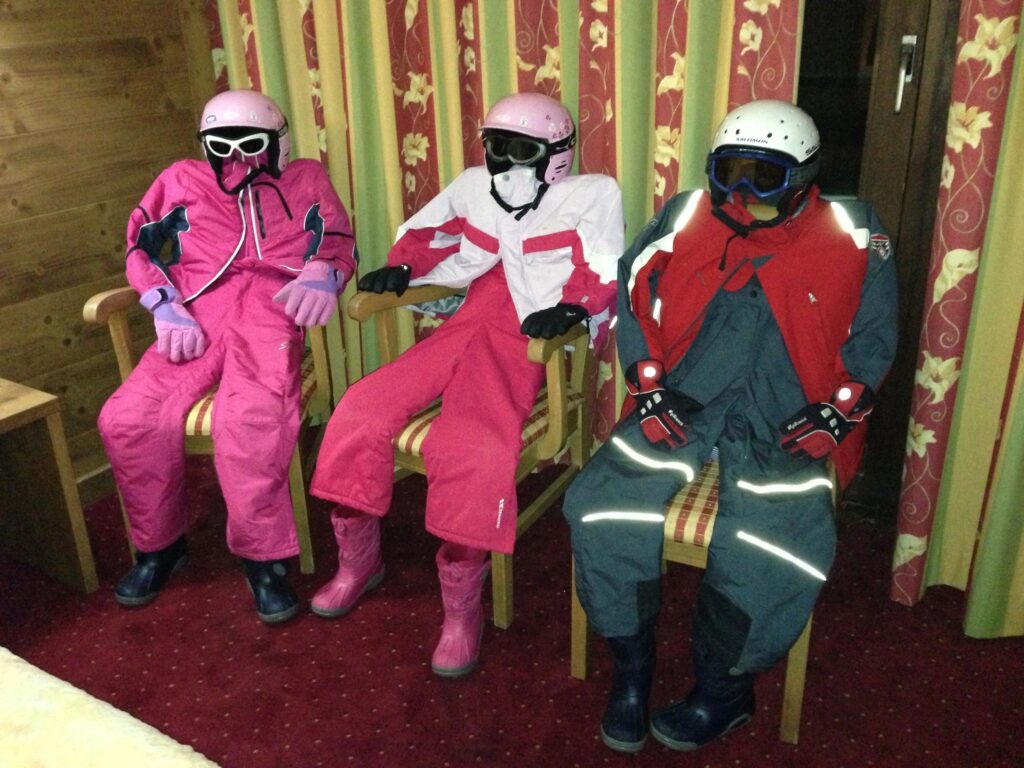 Three figures dressed in ski suits, helmets and goggles seated on chairs